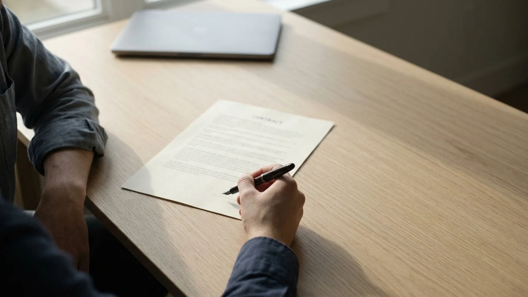 Two people at a wooden desk reviewing a printed contract, one holding a pen above the signature line, daylight from a nearby window